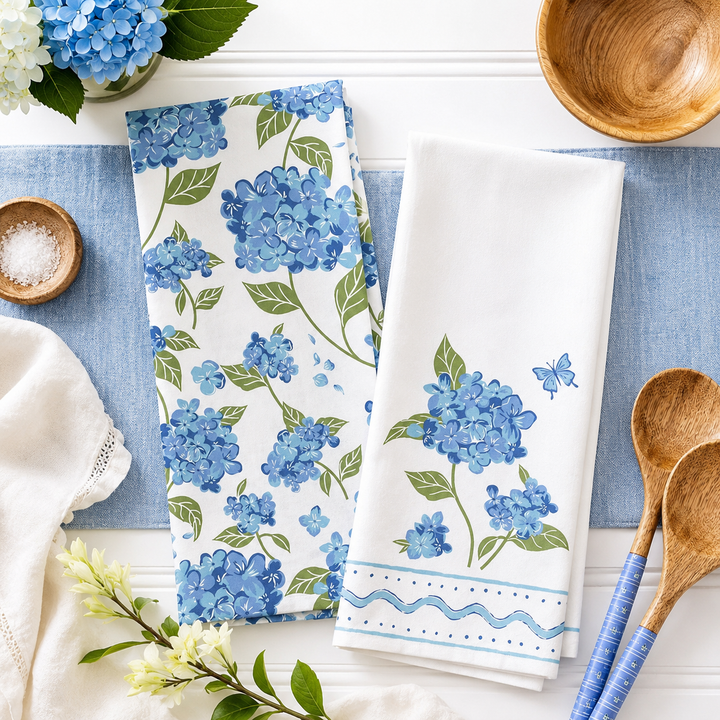 Two white kitchen towels with blue hydrangea floral prints are arranged on a white tabletop with blue placemats and styled props including wooden bowls, spoons, and flowers.