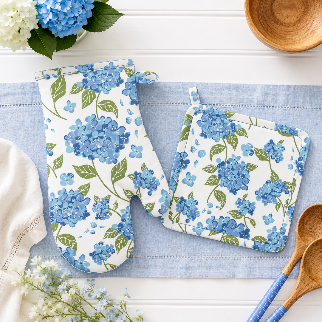 A matching kitchen potholder set with a blue hydrangea floral print on a white background, styled on a light blue runner with wooden spoons, a wooden bowl, and flowers.
